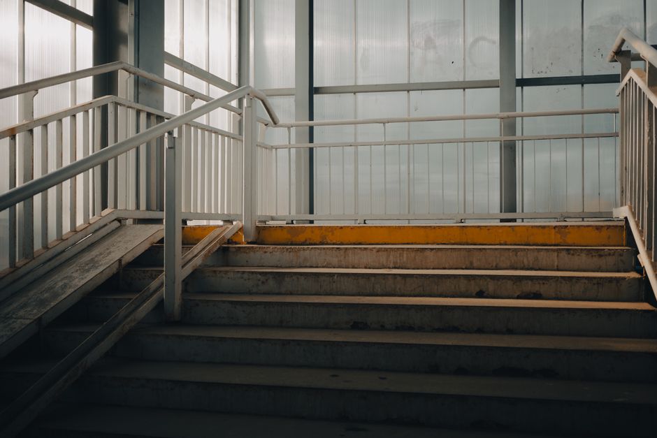 The image shows an indoor staircase with concrete steps that are weathered and stained, leading upwards within a building or industrial setting. The steps are flanked by white metal handrails on both sides, with horizontal bars and vertical supports, appearing clean but with some signs of wear. The environment is illuminated by natural light filtering through translucent panels or windows at the top of the staircase, casting a soft, diffuse glow over the scene. Behind the staircase, there is a backdrop of light gray or metallic wall panels, which may be part of a larger warehouse or industrial structure. The overall atmosphere is neutral and functional, emphasizing the structural elements and the utility of the space, potentially relating to a site where waste or rubbish might be handled or removed, aligning with the themes of private disposal or site clearance that Rubbish Removal Lewisham offers.