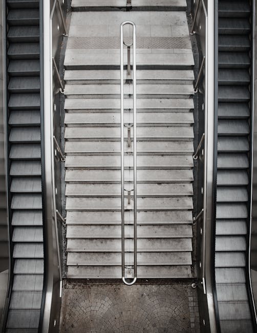 The image shows an indoor staircase with concrete steps that are weathered and stained, leading upwards within a building or industrial setting. The steps are flanked by white metal handrails on both sides, with horizontal bars and vertical supports, appearing clean but with some signs of wear. The environment is illuminated by natural light filtering through translucent panels or windows at the top of the staircase, casting a soft, diffuse glow over the scene. Behind the staircase, there is a backdrop of light gray or metallic wall panels, which may be part of a larger warehouse or industrial structure. The overall atmosphere is neutral and functional, emphasizing the structural elements and the utility of the space, potentially relating to a site where waste or rubbish might be handled or removed, aligning with the themes of private disposal or site clearance that Rubbish Removal Lewisham offers.