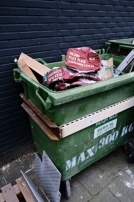 A large green wheeled dumpster filled with red and black bags of garden or building waste, including flexible hoses and packaging, is positioned outside against a dark brick wall. The waste bags display branding and product information, indicating landscaping or construction materials. The container is placed on an outdoor paved surface, with a wooden plank resting on the side and a small metal grate nearby, suggesting a site for on-site waste collection or private rubbish disposal. The scene appears well-lit, capturing the details of the waste and the surroundings, reflecting a typical setting where independent rubbish removal services, such as those offered by Rubbish Removal Lewisham, might be utilized for efficient waste clearance outside residential or commercial properties.