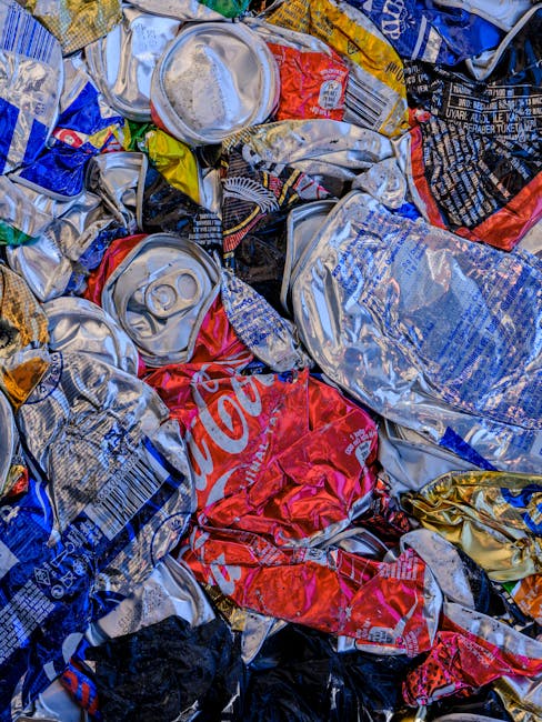 A close-up of a pile of crushed and wrinkled aluminum and plastic beverage cans, predominantly in silver, red, blue, yellow, and black colours. The cans are compressed and overlapping, with some showing visible branding and labels, including portions of a red Coca-Cola can. The surface textures vary from smooth, shiny metal to slightly dented and crinkled areas. The cans are situated outdoors, possibly on a pavement or ground surface, with natural lighting creating reflections on the metal surfaces. The scene suggests a collection ready for disposal or recycling, consistent with waste removal or alternative waste handling services offered by companies like Rubbish Removal Lewisham. The arrangement emphasizes the importance of proper waste segregation and highlights typical recyclable materials managed during private rubbish collection or on-site clearance processes in urban environments.