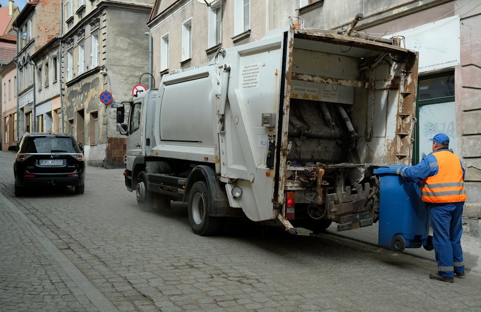 A white waste collection truck with an open rear compartment positioned on a cobblestone street in an urban setting. The truck shows signs of rust and wear on its metal surfaces, which are mainly painted white, with some areas revealing underlying corrosion. A worker dressed in blue overalls with an orange high-visibility vest and blue cap is standing beside a blue wheeled rubbish bin, actively loading or emptying it into the truck's rear compartment. The street is lined with multi-story buildings featuring a mix of old and weathered facades, some with peeling paint and exposed brickwork, and a parked dark-colored car is visible further down the street. The scene is lit with natural daylight, providing clear visibility of the textures and details of the truck, worker, and surrounding environment, illustrating a typical scenario of independent waste collection or rubbish removal service in an urban residential area under the management of Rubbish Removal Lewisham.