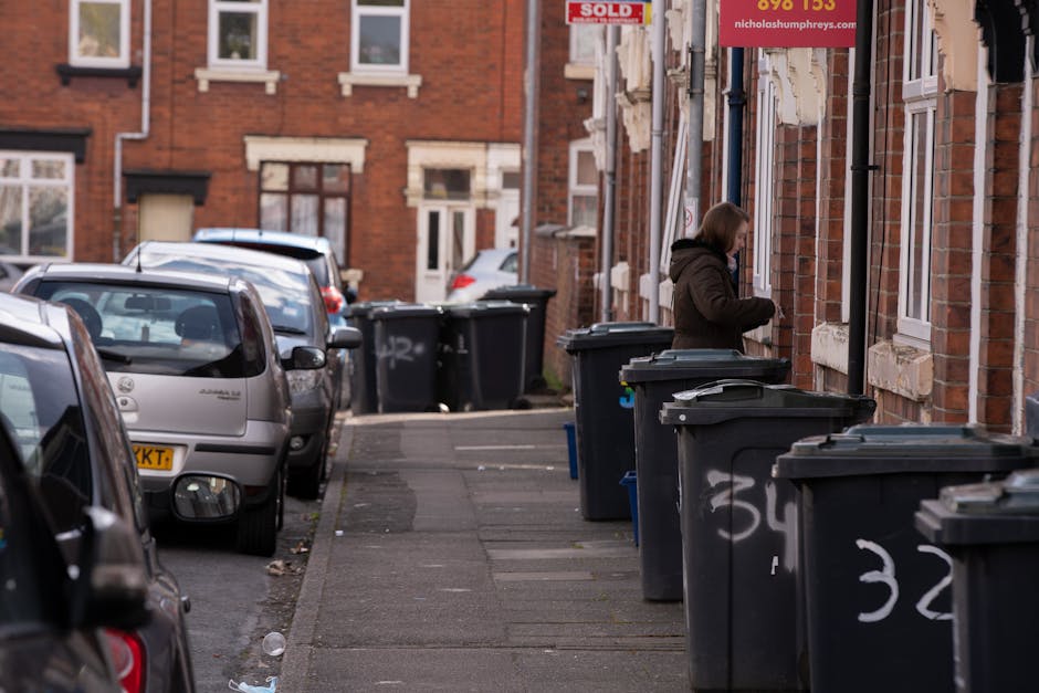 A residential street scene showing a woman standing on the sidewalk next to several large, dark grey wheelie bins lined up along the exterior brick wall of a terraced house. The bins are made of plastic, with some lids closed and others partially open, and each bin has white chalk markings with numbers written on the side. The pavement appears to be concrete, with some areas showing slight discoloration, and there is a narrow curb separating the sidewalk from the parking area where multiple parked cars are visible. Behind the woman, the brick wall features windows with white frames and stone lintels, and a red sign with white text can be seen hanging above. The environment suggests a typical urban setting suitable for private rubbish collection services, with the bins likely awaiting collection or disposal. The lighting indicates daytime, with natural light illuminating the scene evenly, and the overall atmosphere is calm and orderly, reflecting standard rubbish management practices by companies such as Rubbish Removal Lewisham.