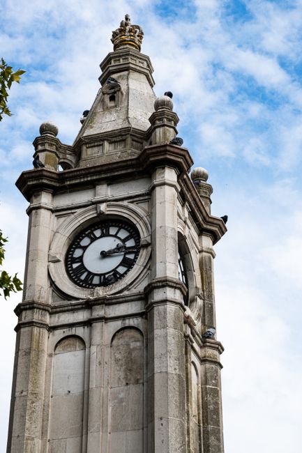 A close-up view of a historic clock tower constructed from light-colored stone, featuring intricate architectural detailing including rounded arches, decorative finials, and small columns. The clock face is prominently displayed with black Roman numerals and hands, showing the time as approximately 9:12. The tower's surface shows weathering and slight discoloration, indicating age, with a clear blue sky dotted with a few wispy clouds in the background. Green foliage from nearby trees partially frames the lower left corner, contrasting with the stone structure. The scene appears to be outdoors, likely situated in a public space such as a town square or park, and while not directly related to rubbish removal, the image's historic architecture emphasizes the importance of maintained communal landmarks. Rubbish Removal Lewisham occasionally handles the clearance of debris around such sites, supporting the preservation of local environment and aesthetics, aligning with alternative waste handling methods through independent collection or on-site clearance services.