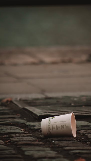 A white disposable coffee cup made of paper with a plastic lid, lying on its side on a cobblestone pavement outside, with a wooden grate or cover nearby. The cup appears empty or discarded, and the surrounding environment includes a blurred background of green and brown tones, suggesting an outdoor setting such as a street or pavement area. The scene captures an instance of littering that could relate to private waste disposal or rubbish removal services in Lewisham, illustrating the importance of responsible waste handling and the role of companies like Rubbish Removal Lewisham in managing such waste efficiently.