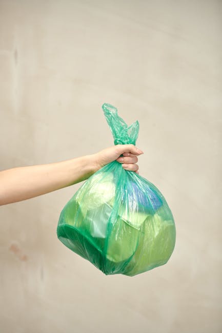 Close-up of several tightly bundled black plastic rubbish bags with glossy, crinkled surfaces, stacked against a plain, light-colored wall in an outdoor or semi-indoor setting. The bags appear full and are secured with twisted tops, with some showing slight creasing and reflections from lighting. They are positioned on or near a surface such as a pavement or driveway, with adjacent bags partially visible in the background, indicating a collection of household waste ready for removal. The environment suggests an area designated for rubbish storage or awaiting collection, aligning with private waste disposal or alternative rubbish removal services like those offered by Rubbish Removal Lewisham. The scene is illuminated by natural or diffuse indoor light, emphasizing the texture and materials of the plastic bags, providing a clear and professional depiction suited for informing about waste management procedures or private collection practices.