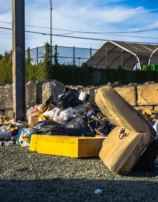 A pile of mixed household waste and rubbish is seen on the ground outdoors, consisting of black plastic trash bags, a yellow plastic storage container, and a discarded beige upholstered armchair with visible fabric texture. The waste is situated on a gravel surface near a stone wall, with some trash spilling over the container and partially covering the armchair. In the background, there is a metal utility pole with power lines, a green hedge, and a large, semi-transparent fabric-covered structure that appears to be a sports or event shelter, set against a partly cloudy sky with patches of blue. The scene suggests an instance of unsorted rubbish awaiting collection, potentially as part of a private waste removal service like Rubbish Removal Lewisham, highlighting the need for efficient waste clearance and disposal solutions.