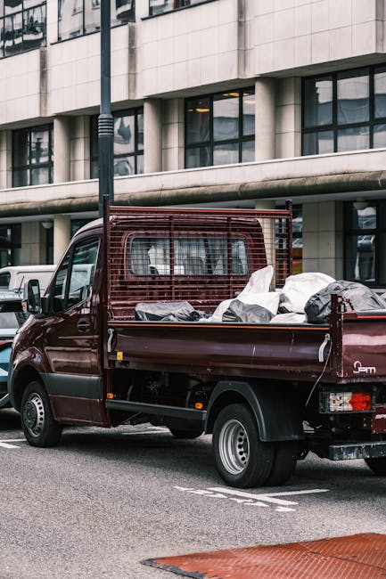 A burgundy flatbed mini truck parked on a city street in front of a modern, multi-storey office building with large glass windows and a concrete facade. The truck's cargo area is loaded with various bags and containers of waste material, including black and white plastic bags, some of which appear to be tied or sealed. The vehicle features a metal mesh safety barrier behind the cab and is positioned on a paved road with visible parking lines. A lamppost stands nearby, and multiple other vehicles are seen parked along the street. The environment suggests an urban setting, potentially involving private rubbish collection or on-site clearance services, as provided by companies like Rubbish Removal Lewisham, operating in the context of alternative waste handling methods outside local authority collections.