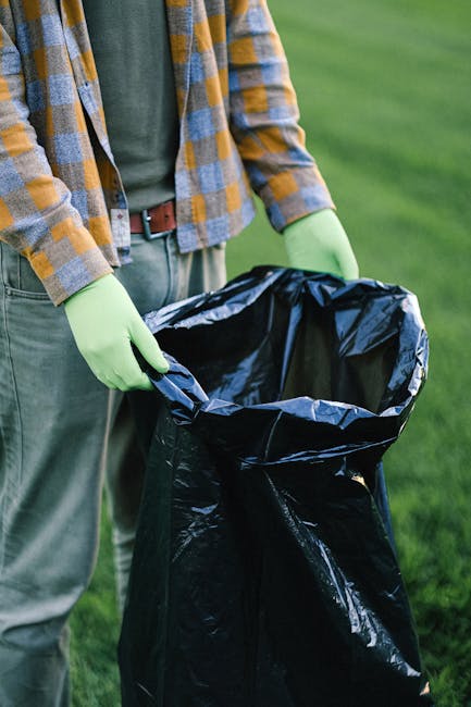 A pile of mixed household waste and rubbish is seen on the ground outdoors, consisting of black plastic trash bags, a yellow plastic storage container, and a discarded beige upholstered armchair with visible fabric texture. The waste is situated on a gravel surface near a stone wall, with some trash spilling over the container and partially covering the armchair. In the background, there is a metal utility pole with power lines, a green hedge, and a large, semi-transparent fabric-covered structure that appears to be a sports or event shelter, set against a partly cloudy sky with patches of blue. The scene suggests an instance of unsorted rubbish awaiting collection, potentially as part of a private waste removal service like Rubbish Removal Lewisham, highlighting the need for efficient waste clearance and disposal solutions.
