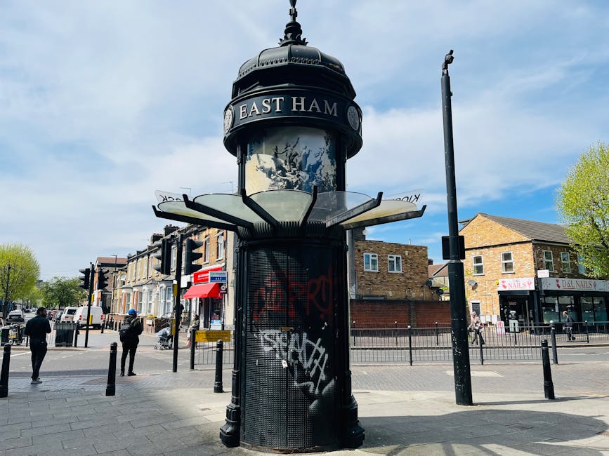 A tall, cylindrical street clock with a domed black metal top and a decorative finial, located at a busy intersection in Lewisham. The clock face is surrounded by a circular band with the text 'EAST HAM,' and the clock itself features large, white numerals and black hour and minute hands against a white background. Below the clock face, there is a mesh-covered vending or information kiosk with graffiti tags in red and white spray paint. The kiosk's finish appears metallic with a black base. The street scene shows pedestrians walking on the paved sidewalk, some wearing dark clothing and hats, with a row of residential-style brick buildings and retail shops in the background, including a nail salon with a pink sign. The area is bordered by black bollards and has a bright sky with some scattered clouds, contributing to a lively urban atmosphere typical of a central Lewisham location with independent councils or public services nearby, subtly tying to the context of performing local waste management or rubbish removal activities in the area. Rubbish Removal Lewisham services are relevant here for maintaining such public spaces through alternative waste handling and onsite clearance solutions.