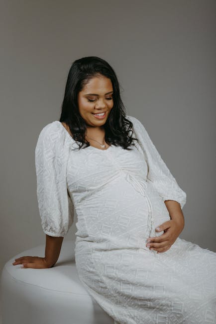 This image features a pregnant woman with medium skin tone and dark, wavy hair sitting on a white surface against a neutral grey background. She is wearing a white textured dress with three-quarter length sleeves, a subtle pattern, and a slightly gathered waist that accentuates her pregnancy. The woman is smiling gently, looking down, with her right hand resting on her lap and her left hand placed on her belly, suggesting a moment of relaxed contemplation. The soft lighting creates minimal shadows, highlighting the smooth finish of the dress and the natural glow of her skin. There are no visible objects or accessories in the background. This natural, artistic portrait emphasizes comfort and maternal tenderness, aligned with themes related to family and care, which can be subtly connected to our waste management services’ focus on accommodating diverse individual needs and environmental considerations.