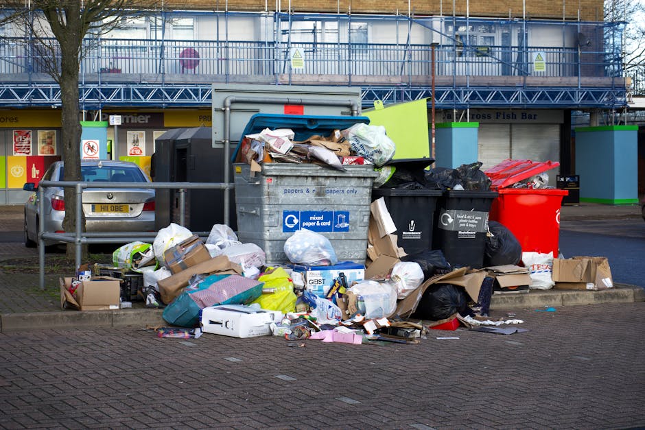 A large collection of mixed household waste and recyclables piled on the pavement in front of a public rubbish collection point, with several black and red rubbish bins, some of which are overflowing with plastic bags and cardboard packaging. The trash includes flattened cardboard boxes, crumpled paper, plastic wrappers, and loose packaging materials, spilling onto the brick surface and surrounding the bins. Behind the waste disposal area, a metal railing separates the rubbish zone from a parked silver car with a yellow registration plate. In the background, there are glazed shopfronts with signs, and above them, a blue metal scaffolding and a temporary construction barrier partially obscuring a building under renovation. The scene is outdoors under overcast lighting, illustrating an example of improper waste disposal that local rubbish removal services, like Rubbish Removal Lewisham, can help manage through efficient collection and clearance solutions.