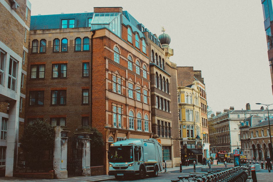 A street scene in an urban area featuring a variety of multi-storey buildings with diverse architectural styles, including brick facades and ornate window framing. In the foreground, a white waste collection vehicle with a blue and black logo on the cab is parked on the road, positioned to the left of the image. The vehicle appears to be part of private rubbish removal services and is parked close to a row of metal bike racks along the curb. To the right, a narrow sidewalk runs alongside the buildings, with a series of black metal bollards separating pedestrian space from the road. The sky is overcast, creating diffuse natural lighting that illuminates the scene evenly. The environment suggests an active city centre with nearby shops and mixed-use buildings, aligning with the context of independent waste collection and on-site clearance managed by local firms such as Rubbish Removal Lewisham. The overall scene emphasizes urban rubbish removal operations within a dense, historic commercial district in Lewisham.