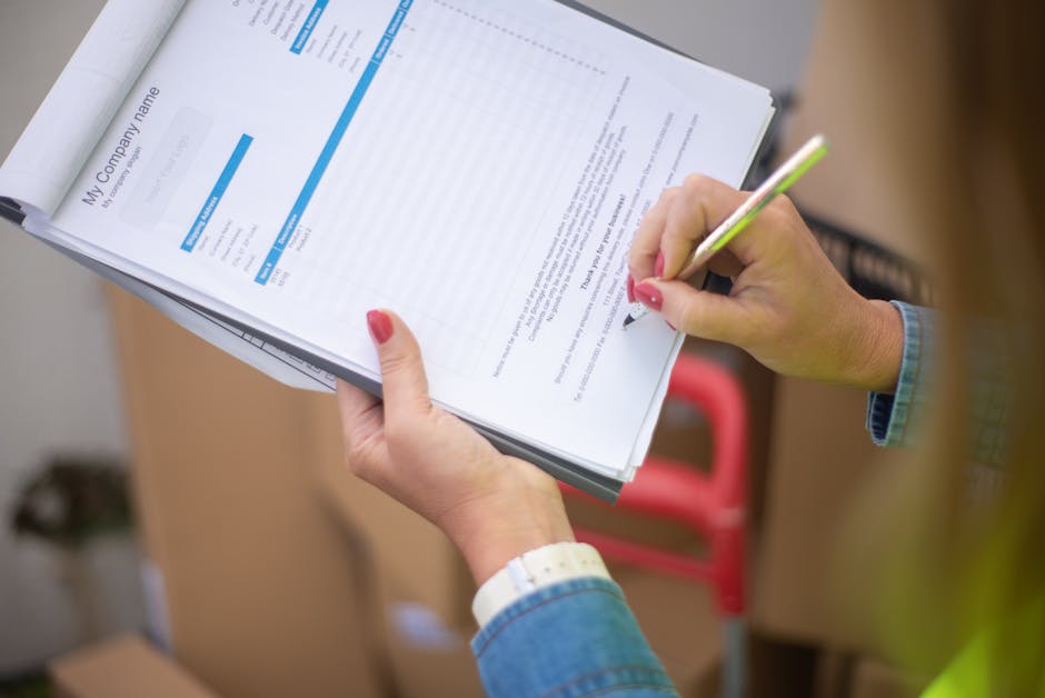 A person with neatly manicured nails painted red is holding a clipboard with multiple sheets of paper, which include a document titled 'My Company Name' with blue and white design elements, in an indoor setting. The individual is using a pencil with a green tip to make notes or fill out the form. The person is wearing a denim jacket, and a white wristwatch is visible on their left wrist. The background shows a blurred view of a workspace, with part of a red handle or tool, and some cardboard boxes, suggesting an environment related to office work or logistical tasks. This scene may relate to administrative procedures involved in rubbish removal or waste management, such as completing transfer notes or licensing documentation, typical of services like those offered by Rubbish Removal Lewisham, which provide independent waste handling and disposal services in Lewisham.